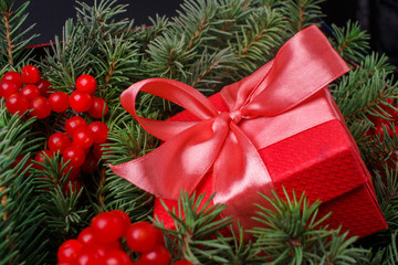 New Year Christmas Xmas 2020 holiday celebration red present gift box with satin pink bow, immersed in the needles of a Christmas tree decorated with red berries. Dark festive composition.