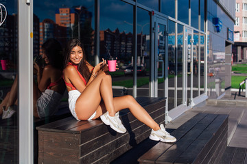 Stylish happy young brunette woman wearing white shorts and sneakers holding pink cup of coffee to go sitting next to coffee shop.