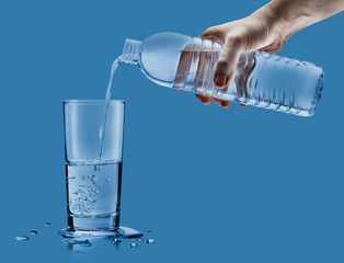Hand pouring mineral water from bottle into a glass with water drops in the blue background,...