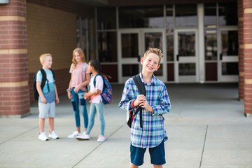 Handsome pre-adolescent teen boy student hanging out with friends after school. Selective focus on the smiling boy student standing outside the school building with friends in the background - obrazy, fototapety, plakaty