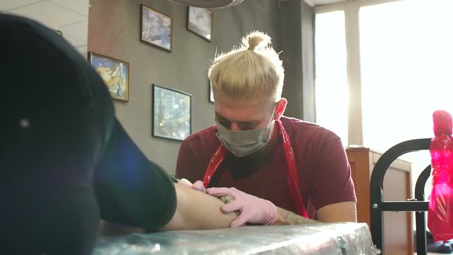 Portrait of a modern tattoo artist while working with a client. A professional tattoo artist makes a tattoo on the leg of a young girl in a tattoo parlor. Slow motion.
