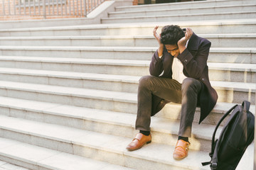 Young handsome Indian man in a white shirt and a business jacket. Sitting sad on the steps of the building with his hands clasped. The concept of dismissal, layoffs, unemployment. University Exam