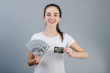 beautiful young woman smiling and holding one hundred dollar bills and credit card isolated over grey