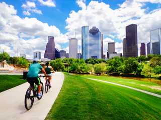 Riding Bikes on Paved Trail in Houston Park (view of river and skyline of downtown Houston) - Houston, Texas, USA  - obrazy, fototapety, plakaty