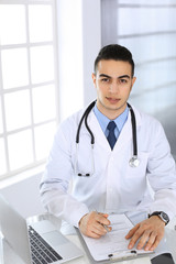 Arab doctor man using laptop computer while filling up medication history records form at the glass desk in medical office or clinic. Medicine and healthcare concept