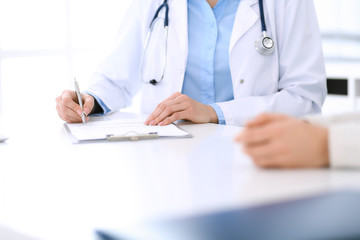 Woman doctor and patient sitting and talking at medical examination at hospital office, close-up. Physician filling up medication history records. Medicine and healthcare concept