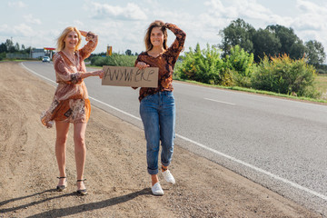 Two beautiful girls hitchhiking and vote with a sign ANYWHERE on road. Copy space.