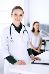 Doctor woman at work with patient and colleague at background. Physician filling up medical documents or prescription while standing in hospital reception desk. Data in medicine and health care