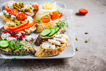 Open toasts with different toppings on gray-brown background. Crostini with banana, pate, avocado, salmon, egg, cheese and berries.