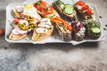 Open toasts with different toppings on gray-brown background. Crostini with banana, pate, avocado, salmon, egg, cheese and berries.