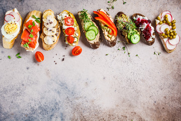 Open toasts with different toppings on gray-brown background, copy space, top view.  Flat lay of crostini with banana, pate, avocado, salmon, egg, cheese and berries.