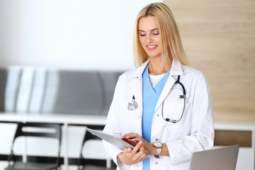 Doctor woman at work in hospital excited and happy of her profession. Blonde physician controls medication history records and exam results while using tablet computer. Medicine and healthcare concept