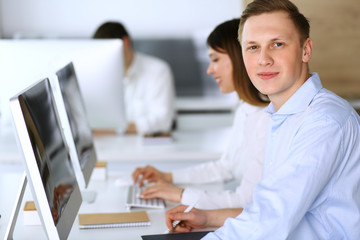 Cheerful smiling businessman headshot at work in modern office. Unknown casual dressed entrepreneur using pc computer while sitting with diverse colleagues at the background. Multi ethnic working