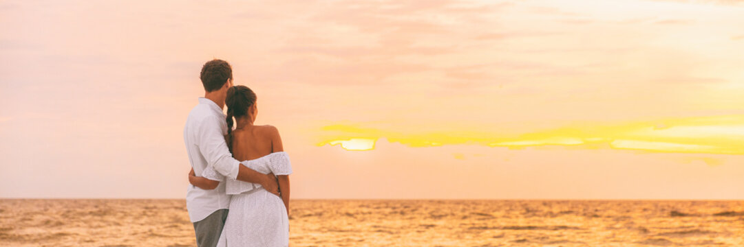 Honeymoon couple walking on romantic evening watching sunset banner panorama background. Beach wedding woman wearing white dress and elegant man lovers relaxing on Caribbean vacation travel panoramic.