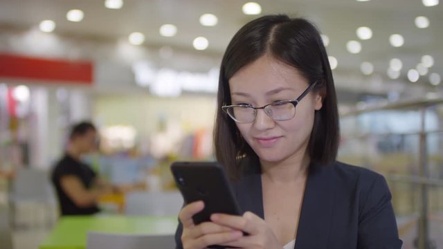 Chest-up arc shot of young Chinese woman in glasses sitting in shopping mall, looking at smartphone screen with happy smile, and typing message