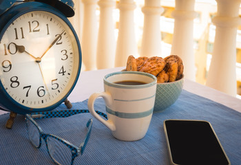 A small table outside on the balcony with a cup of coffee and some biscuits with chocolate for a break or breakfast. Old blue metal alarm clock. Mobile phone and eyeglasses