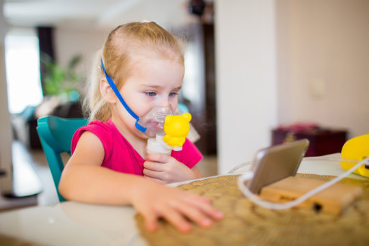 Little girl with allergic asthma using inhaler and watching cartoons on a smartphone. Girl inhales medicine through a nebulizer mask. Treatment of the respiratory tract.