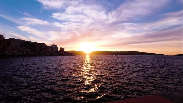 A timelapse sunset wide shot taken in St. Paul's Bay, San Pawl il-Bahar, Gillieru Harbour, Malta in early 2019. Golden hour to dusk, calm sea with reflections. Street Lights appear.