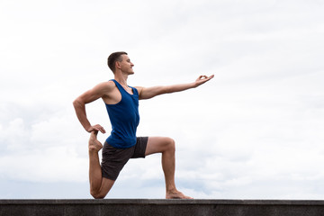 Obraz na płótnie Canvas smiling young man is doing yoga in city at summer time against sky background