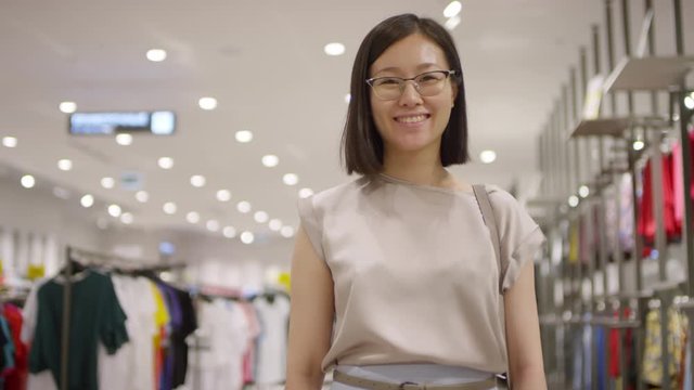 Waist-up arc shot of smiling young Chinese woman, wearing beige top and glasses, standing in clothing store in front of racks and rails and looking at camera