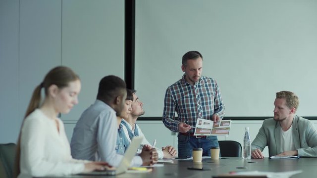 Tracking shot of diverse business team discussing application design in meeting. Man presenting interface templates on paper to colleagues in board room