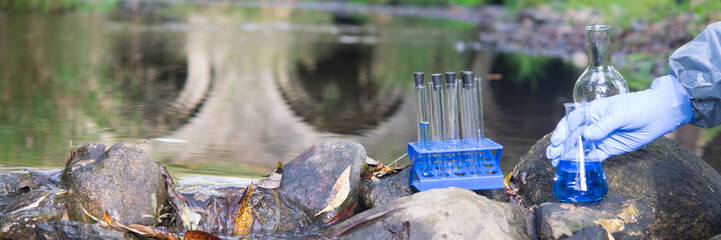 scientist's hand, in a protective suit and gloves, collects liquid into flasks and test tubes, for testing, against the background of the river
