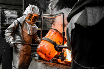 In a foundry workshop. A worker protected by a safety suit pours the molten metal into a mold - 298807791
