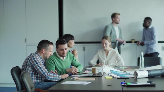Tracking shot of diverse business people working in office meeting room. Two men discussing documents at desk, female colleagues making notes in notebook nearby. Two businessmen talking in background