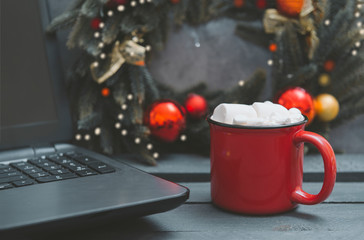 mug of cocoa with marshmallows next to a laptop, a Christmas wreath with Golden and red balls on a gray background,