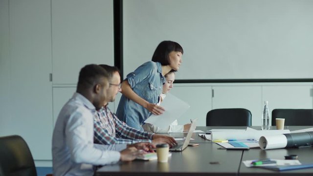 Group of diverse business people gathering at table in meeting room. Middle aged professional presenting website on laptop and smartphone screens to colleagues, tracking shot