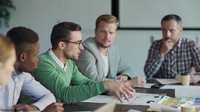 Tracking shot of diverse team of business people discussing market analysis sitting at desk in meeting. Intelligent young man in eyeglasses explaining statistics to colleagues