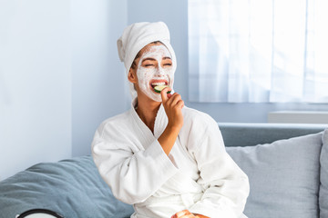 Beautiful young woman with facial mask on her face holding slices of fresh cucumber. Skin care and treatment, spa, natural beauty and cosmetology concept
