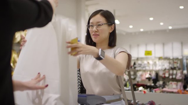 Waist-up shot of cheerful young Chinese woman paying with credit card at checkout counter of clothing store, taking shopping bags from sales assistant, thanking her and leaving