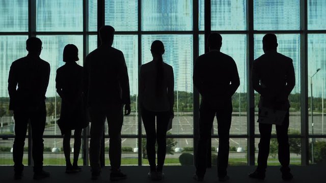 Rear view backlit full length shot of silhouettes of six business people standing by panoramic window with documents in their hands, looking at future building site and having discussion