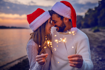 Overjoyed caucasian couple in love standing on coast near river and holding sparklers. Both are dressed in white sweaters and having santa hats on heads. - Powered by Adobe