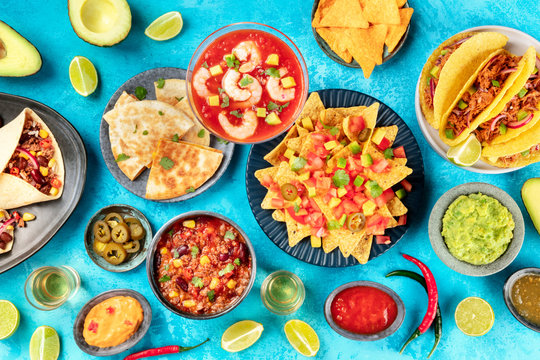 Mexican Food, Many Dishes Of The Cuisine Of Mexico, Flat Lay Overhead Shot From On A Vibrant Blue Background. Nachos, Tequila, Guacamole, Chili Con Carne, Burritos, Tacos