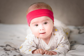 Adorable 6 months old Baby girl infant on a bed on her belly with head up looking into camera with her big eyes. Natural light.