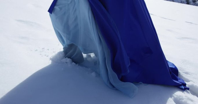 Woman in blue dress is walking through snowland. Footprints on the snow and hem clouseup.