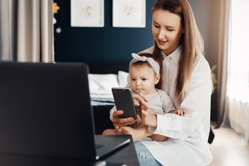 Mother with baby daughter sitting in front of laptop and watching videos and photos on smartphone. Entertaining content for children on mobile phones, educating games, interesting cartoons, learning.