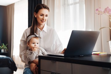 Female freelance author typing on laptop and holding baby on knees. Vacancies for home-based remote...