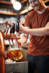 Handsome caucasian barman pouring beer while standing in pub. Selective focus on hand.