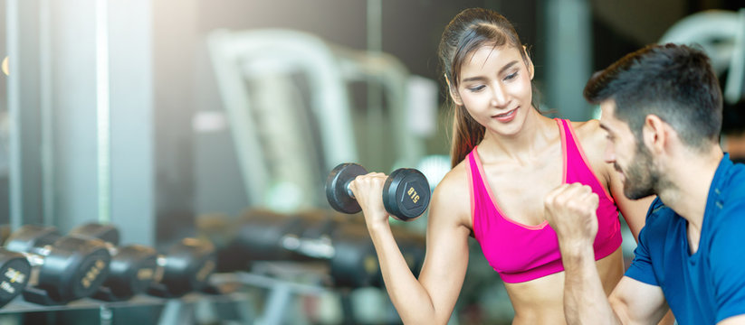 Banner of Trainer man is teaching asian spoty woman lifting a dumbbell and exercise in the gym. Smiling Woman Using Hand Weights While Personal Trainer Supervises Her Progress