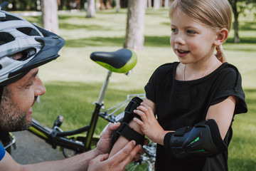 Obraz premium Young man in protective helmet helping daughter to put on elbow pads