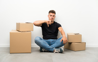 Handsome young man moving in new home among boxes showing thumb down sign
