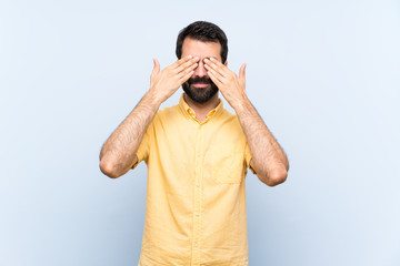 Young man with beard over isolated blue background covering eyes by hands