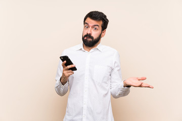 Young man with beard holding a mobile having doubts while raising hands