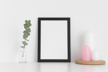 Black frame mockup with candles and eucalyptus in a vase on a white table. Portrait orientation.