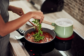 Mixed race woman in apron standing next to stove, stirring tomato sauce and adding rocket. On stove is pot with boiling pasta.