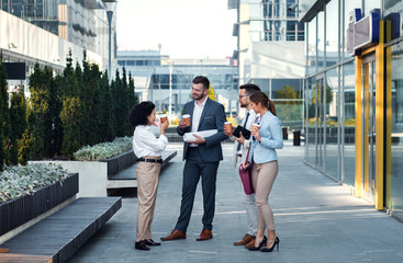 Group of coworkers having a coffee brake together, standing outside in front of office buildings and talking.