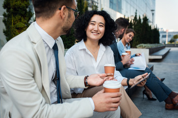 Group of coworkers having a coffee brake together, siting outside in front of office buildings and talking.